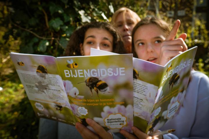 2 meisjes en een dame staan buiten achter het bijengidsje. het rechter meisje wijst naar iets buiten het scherm, de andere twee personen kijken naar de gids